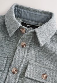 Veste-chemise à chevrons gris clair, dotée d'une fermeture à boutons, de deux poches poitrine et d'un col arrondi. L'étiquette indique "NEXT, Fabriqué en Birmanie."