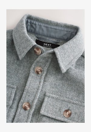 Világosszürke halszálka mintás ingdzseki, gombos elülső résszel, két mellkasi zsebbel és lekerekített gallérral. A címke felirata: "NEXT, Made in Myanmar."