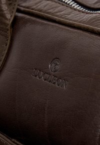 Brun lædertaske med en glat tekstur, der har et debossed logo og en lynlås lukning. Understreger kvalitetshåndværk og design detaljer.