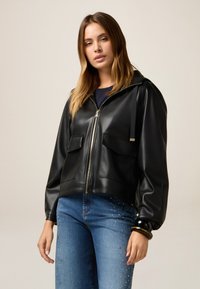 Zwarte neplederen jas met een capuchon, rits aan de voorkant, twee grote zakken en elastische manchetten, gecombineerd met blauwe jeans met versieringen.
