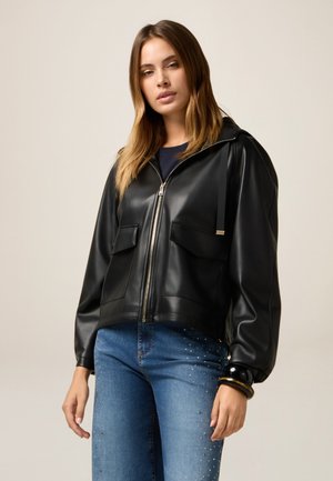 Zwarte neplederen jas met een capuchon, rits aan de voorkant, twee grote zakken en elastische manchetten, gecombineerd met blauwe jeans met versieringen.