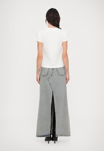 Džínová maxi sukňa svetlošedej farby má vysoký rozparok, zadné vrecká a kontrastné švy. Kombinovaná s bielym tričkom s krátkym rukávom.