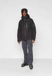 Black ski jacket with front zippers, multiple pockets, and adjustable hood. Paired with gray snow pants and black ski boots.