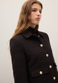 Jonge vrouw met lang golvend haar draagt een donkerbruin geruit jasje met gouden knopen en een donkerbruine coltrui, kijkt naar rechts.