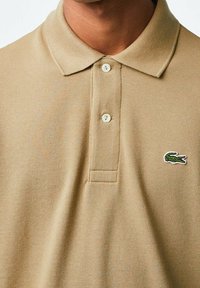 Polo beige en tissu texturé, doté d'un col et de trois boutons. Le logo vert en forme de crocodile est bien visible sur le côté gauche.