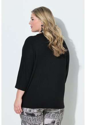 Donna con i capelli biondi lunghi, indossa una maglietta nera e pantaloni a fantasia, rivolta verso sinistra su sfondo bianco.