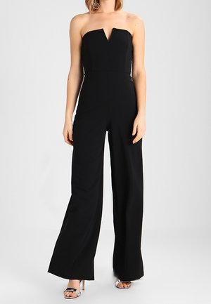 Žena nosící černý široké kalhoty bez ramínek s pevně tvarovaným korzetovým vrškem a stříbrné sandály na vysokém podpatku, stojící před jednobarevným pozadím.