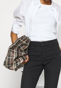 Jean noir taille haute associé à un débardeur blanc côtelé, sous une chemise blanche ouverte à boutons ; tenant un flanelle à carreaux marron.