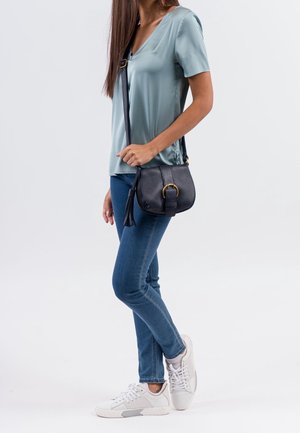 Sac bandoulière en cuir noir avec une finition texturée, des ferrures dorées et un accent de pampille. Porté avec un haut bleu sarcelle clair et un jean bleu.