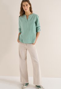 Femme debout, les mains dans les poches, portant un chemisier texturé vert clair à manches longues, un pantalon large beige et des baskets blanches avec des détails verts.