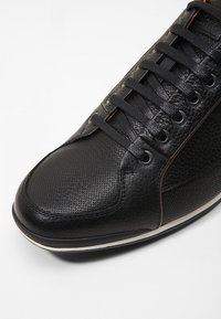 Baskets en cuir noir avec orteil perforé, côtés texturés, accents marron contrastants, et lacets plats noirs avec œillets ronds.