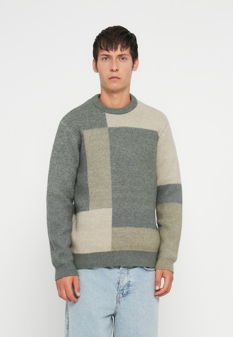 Pull en tricot gris et beige avec un motif blocs, doté d'un col rond et de poignets côtelés. Texture douce ; coupe décontractée.