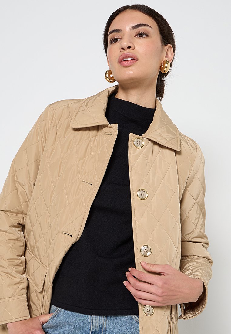 Donna che indossa una giacca trapuntata beige sopra un top nero e jeans blu, con orecchini a cerchio dorati, in posa contro uno sfondo bianco.