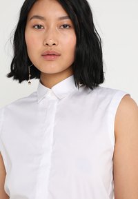 Jeune femme aux cheveux noirs mi-longs portant une chemise blanche sans manches boutonnée et une boucle d'oreille en or pendante à une oreille.