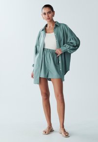 Světle zelená oversized košile na knoflíky spárovaná s odpovídajícími šortkami. Košile má nařasené rukávy a outfit doplňuje bílý žebrovaný top bez rukávů.