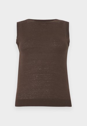 Femme debout, main dans la poche, portant un haut marron sans manches et un pantalon noir large contre un fond gris uni.