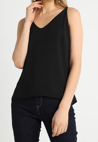 Top negro sin mangas con escote en V, hecho de tela suave. Presenta un ajuste relajado y sutiles aberturas laterales, combinado con jeans de mezclilla oscura.