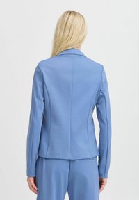 Blazer bleu clair en tissu texturé, présentant une coupe structurée, des manches longues et un col cranté, avec des détails de couture visibles.