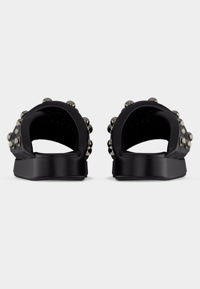 Sandales noires à enfiler avec de larges brides ornées de clous ronds métalliques, vues de dos sur un fond blanc.