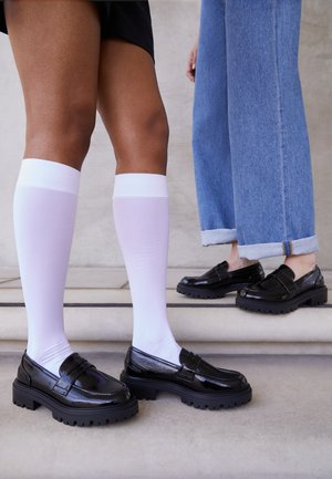 Černé lakované kožené mokasíny s masivními černými podrážkami, v kombinaci s bíložitými punčochami po kolena; jedna osoba v modrých džínách, druhá v černé sukni.