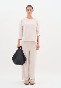 Un sweat-shirt à manches longues beige clair, assorti à un pantalon large beige et des chaussures à enfiler beiges. Un sac à main en cuir noir est tenu dans la main droite.