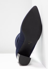 Chaussure bleu marine à bout pointu avec semelle noire texturée et talon bloc, taille 37, fabriquée en Espagne, inclinée contre une surface blanche d’angle.
