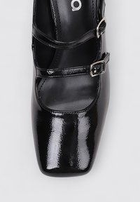 Zapato de charol negro con punta cuadrada, tiras cruzadas y detalle de hebilla plateada. Presenta un acabado suave y brillante.