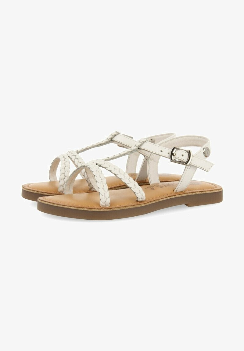 Sandales plates blanches avec brides tressées, boucle à la cheville, semelle intérieure marron clair et semelle extérieure brune, vues de côté sur un fond blanc.
