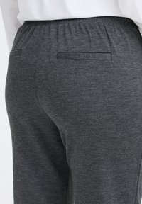 Gros plan sur un pantalon gris foncé avec une taille élastique et deux poches passepoilées à l'arrière, porté avec une chemise blanche.