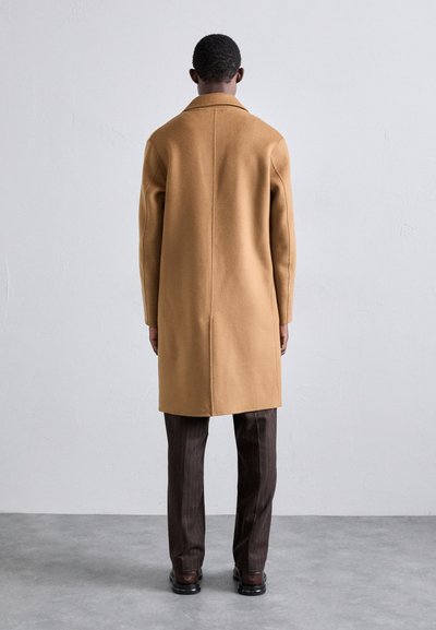 Tan wollen overcoat met een getailleerde pasvorm, voorzien van een rechte snit en lange mouwen, in combinatie met bruine pinstripe broeken en zwarte schoenen.