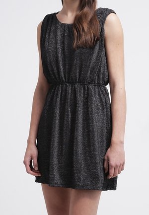 Vestido negro sin mangas con brillo y cintura fruncida, llevado por una persona que está de pie contra un fondo claro y liso.