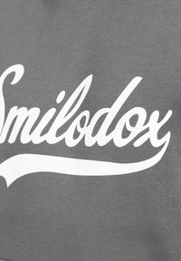 Szara tkanina o gładkiej fakturze z białym kursywą napisem "Smilodox" oraz zakrzywioną linią akcentującą poniżej.