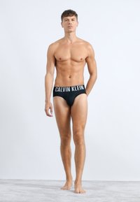 Čierne klasické nohavičky Calvin Klein s bielym logom. Model stojaci naboso na svetlošedej ploche, predvádzajúci strih a dizajn.