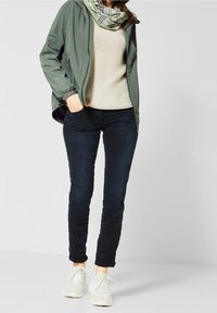 Olivově zelená lehká bunda, béžový pletený svetr, tmavě modré džíny a bílé tenisky. Šála má károvaný vzor se zelenými akcenty.