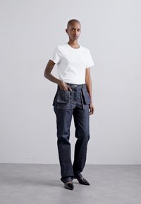 Valkoinen lyhythihainen T-paita, jossa on teksti, yhdistettynä tummiin farkkuhousuihin, joissa on suuret etutaskut ja löysä istuvuus. Terävät mustat kengät.