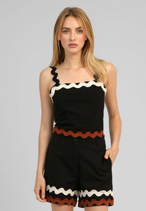Femme blonde portant un haut noir sans manches et un short avec bordure ondulée blanche et marron, debout, une main dans la poche, sur fond gris.