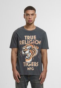 Kokogai pelēks kokvilnas t-krekls ar tīģera grafiku oranžā un baltā krāsā, zem kura ir uzraksts "TRUE RELIGION TIGERS MFG". Īsās piedurknes.