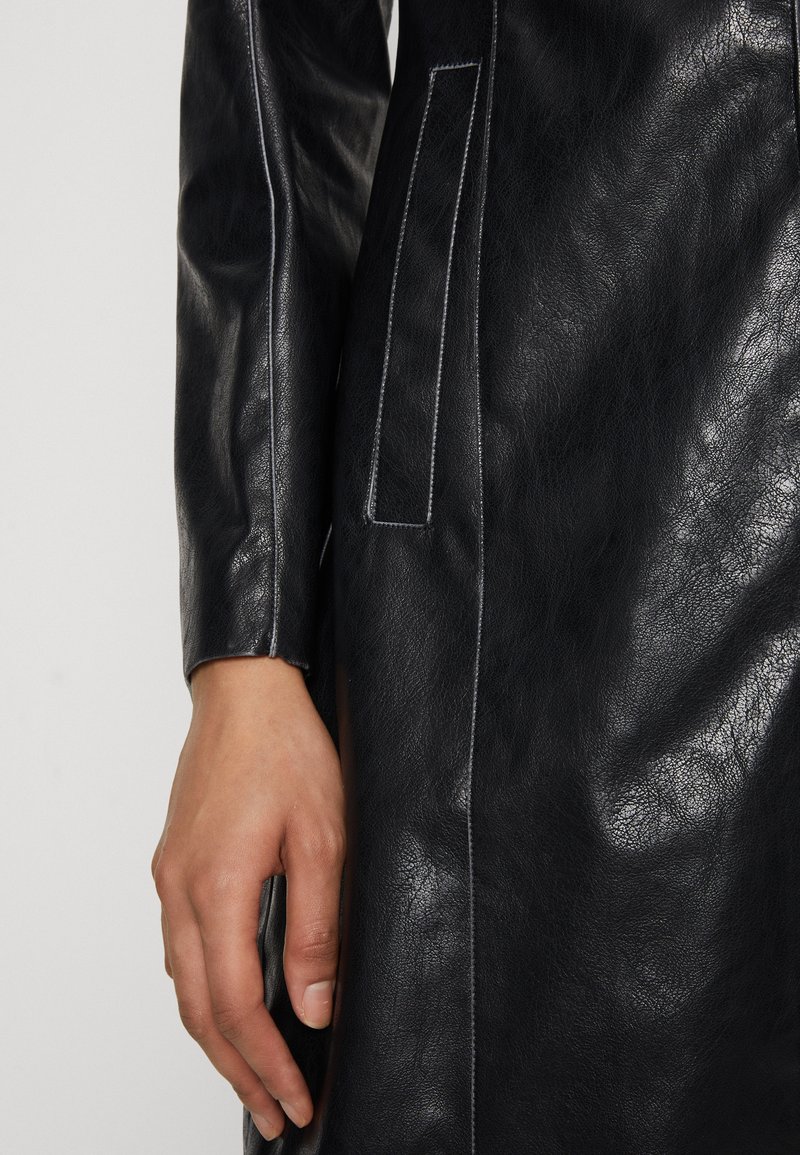 Abrigo de cuero negro con una textura suave, que presenta costuras en blanco en contraste y un bolsillo lateral. Primer plano de la manga y la mano.