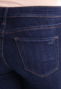 Mörkblå denimjeans med en bakficka med orange sömnad och en liten logotyp. Tyget har en slät, mjuk struktur.