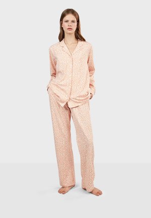 Femme debout pieds nus portant un haut boutonné à manches longues rose clair et un pantalon ample assorti avec un petit motif léopard.