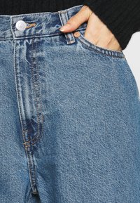 Vaaleansiniset farkut, joissa on korkea vyötärö, kuparinväriset yksityiskohdat, kontrastivärinen ompelu ja tasku oikealla puolella.