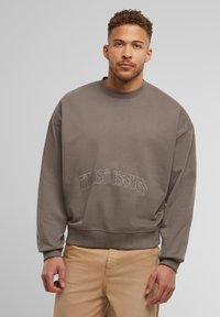 Brązowy sweter z okrągłym dekoltem i haftowanym napisem "TWIST ISSUES". Posiada luźny krój oraz ściągacze na rękawach. W zestawie z beżowymi spodniami.