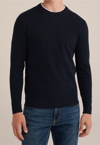 Man som bär en mörk marinblå strukturerad stickad tröja över en vit skjorta, kombinerad med mellanblå jeans, står mot en beige bakgrund.