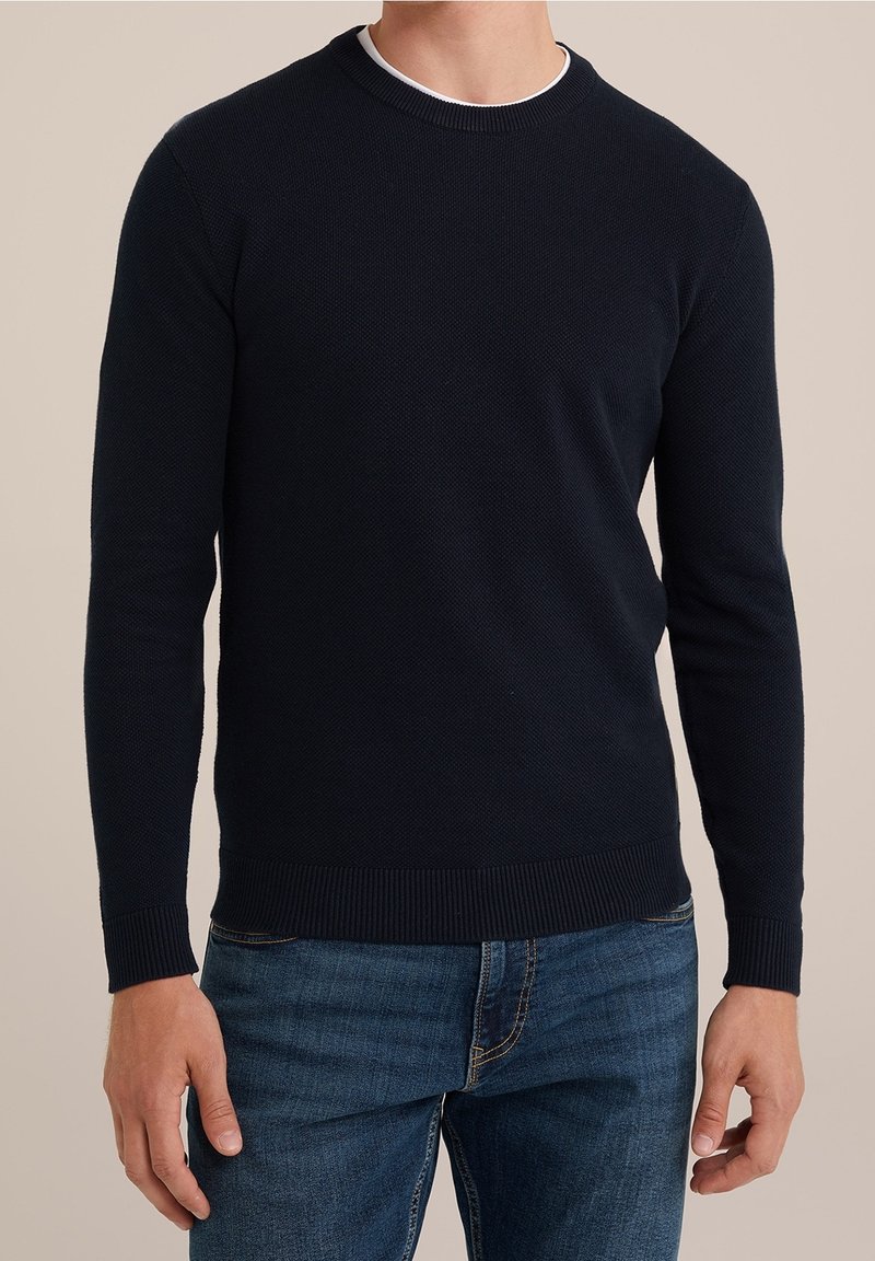 Man som bär en mörk marinblå strukturerad stickad tröja över en vit skjorta, kombinerad med mellanblå jeans, står mot en beige bakgrund.