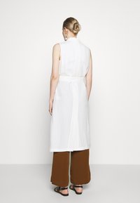 Weißes ärmelloses Kleid mit einem taillierten Gürtel, geradem Schnitt und Schlitz hinten, kombiniert mit braunen weit geschnittenen Hosen und schwarzen Sandalen.