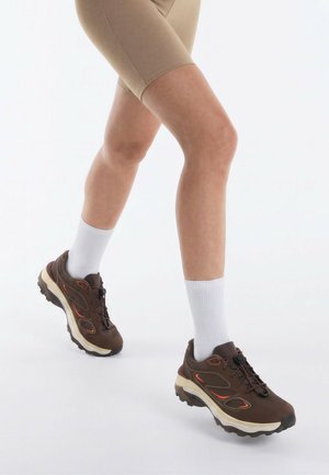 Gambe con pantaloncini beige, calzini bianchi e scarpe da trekking marroni che fanno un passo avanti su uno sfondo bianco uniforme.