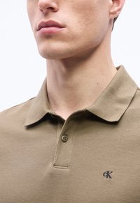 Νεαρός άνδρας που φοράει μπεζ πόλο με γιακά και μικρό λογότυπο "cK" στο αριστερό μέρος του στήθους, εμφανιζόμενος από τον λαιμό έως το άνω μέρος του στήθους.