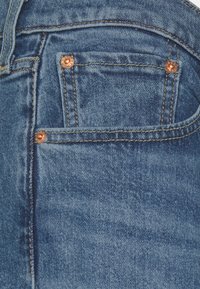 Denimjeans med en medelblå tvätt, prydda med kopparnitar, kontrastsytt och en liten framficka. Slät textur och klassisk design.