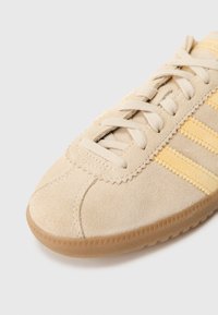 Μπεζ πάνινο παπούτσι από σουέτ με κίτρινες ρίγες και ανοιχτά καφέ λαστιχένια σόλα, με διασταυρωμένα κορδόνι�α σε λευκό φόντο.