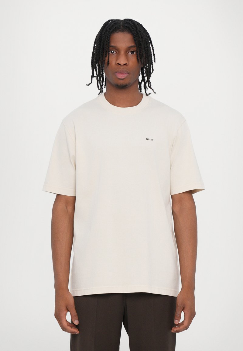 Camiseta beige de algodón, con cuello redondo y mangas cortas. Pequeño texto negro "NN. 07" en el lado izquierdo del pecho. Diseño simple y minimalista.